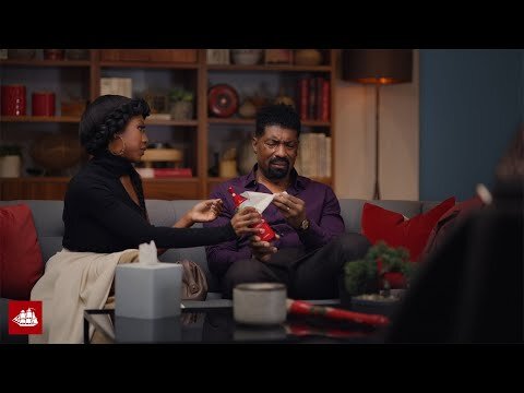 An African American man and woman sit on a couch with a box of tissues and cleaning spray in a modern living room setting.