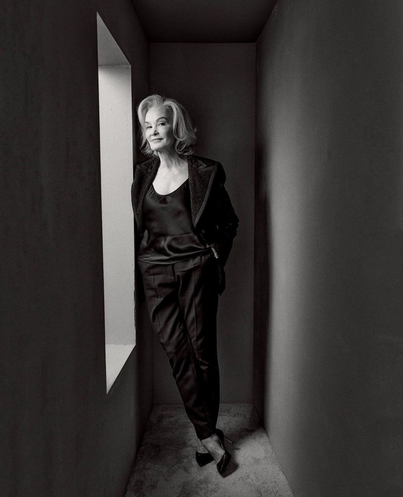 Elegant mature woman poses in a black satin top and suit pants in a modern interior with a narrow window and neutral colors.
