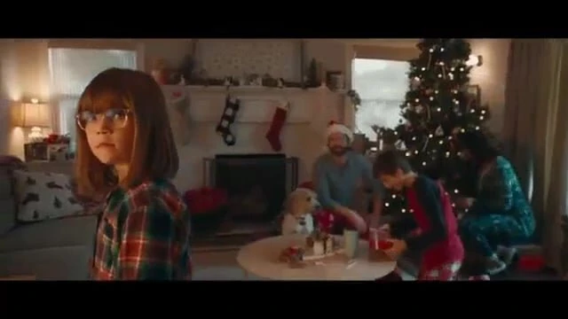 A family of five, including a child in glasses, gathers in a warmly lit living room around a Christmas tree, celebrating the holiday.