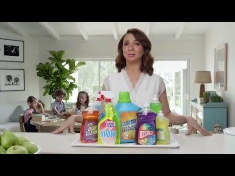 Woman stands behind an array of colorful cleaning product bottles on a white counter with children at a table in the background.