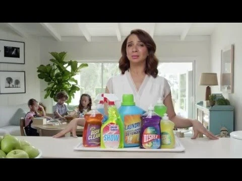 Woman stands behind an array of colorful cleaning product bottles on a white counter with children at a table in the background.