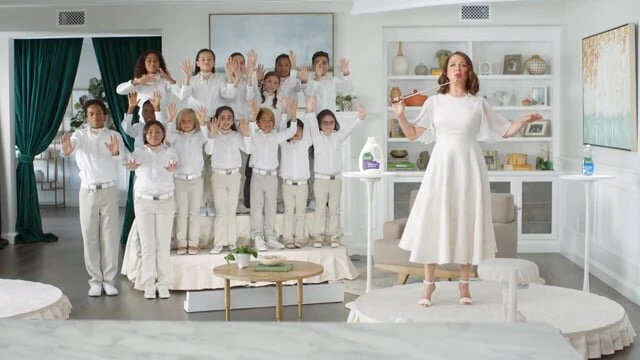 A woman in a white dress conducts a multi-ethnic group of children in white shirts and pants, set against a bright, minimalist living room backdrop.