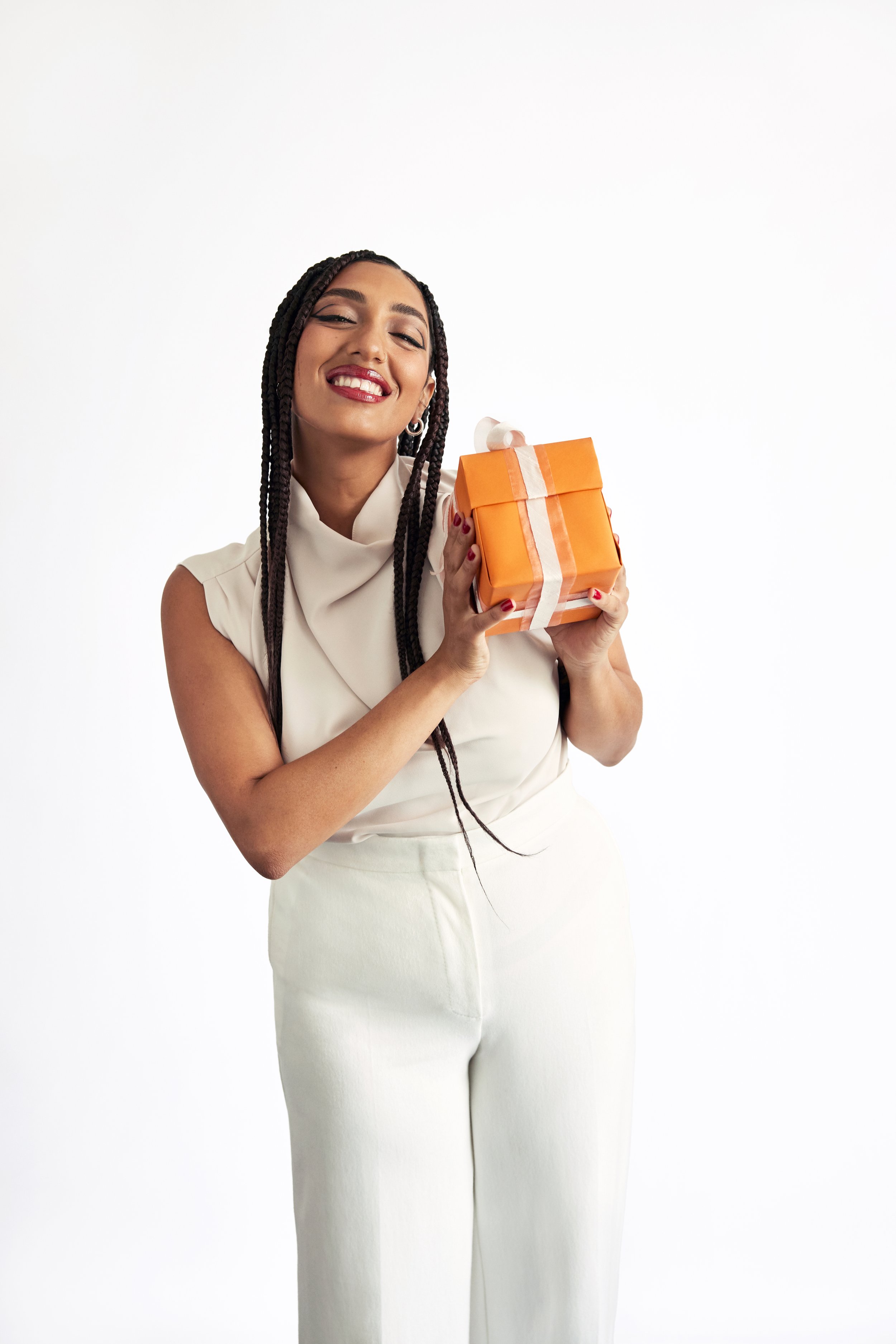 Beautiful adult female smiles while holding an orange gift box with a white ribbon in a studio setting on a white backdrop.