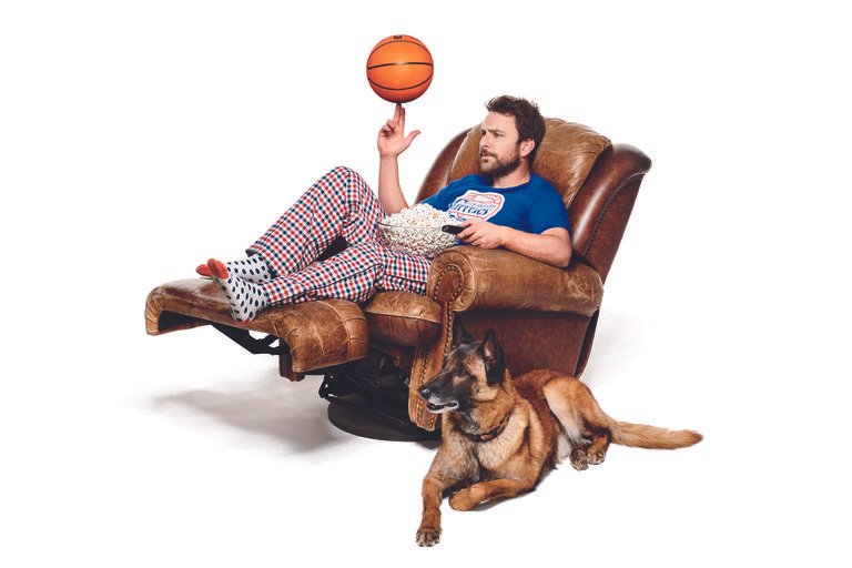 Man in pajamas sits in leather recliner, spinning basketball on finger, dog nearby. He holds popcorn and a remote control.