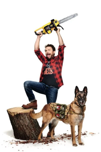 Man in flannel shirt poses with chainsaw above head next to Belgian Malinois dog wearing tactical pack in a white studio setting.