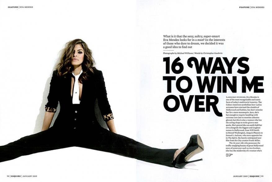 Eva Mendes poses in a black pantsuit on a white background for a magazine cover, sitting with her legs spread. 