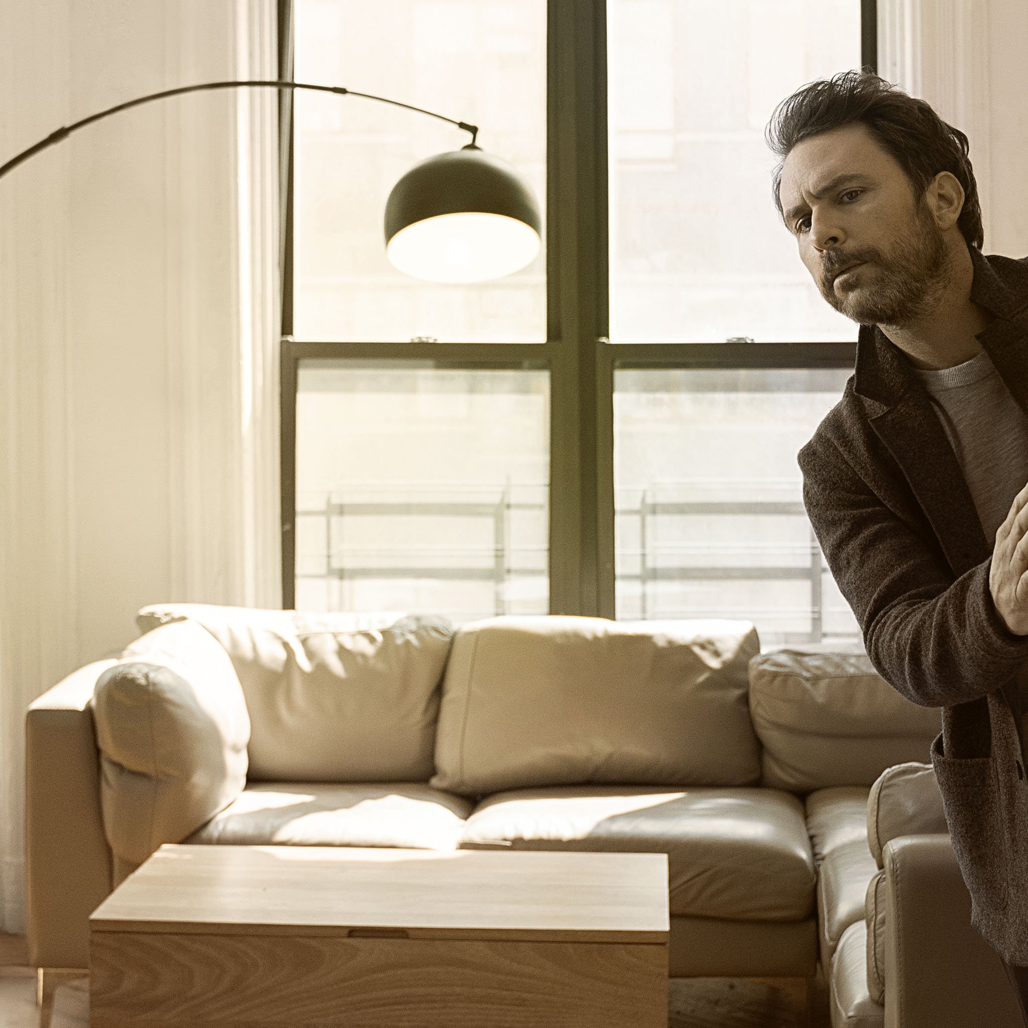 Indoor view of a man looking sideways from behind a cream sofa next to a wood coffee table, and modern black floor lamp near a window.