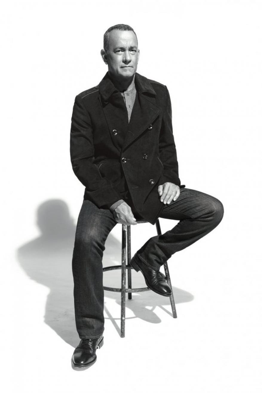 Black and white portrait of adult male in peacoat and jeans, sitting on metal stool in photo studio, looking directly at camera.