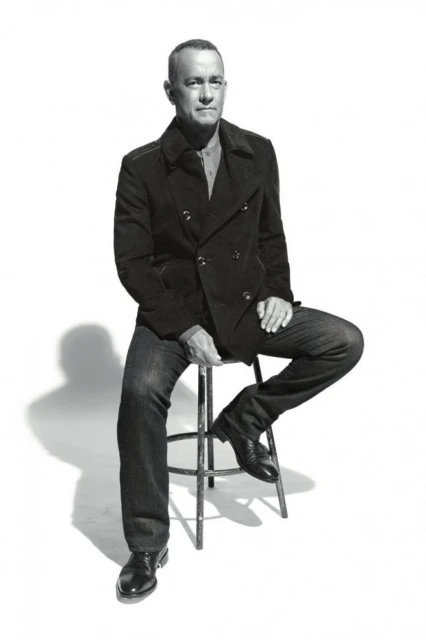 Black and white portrait of adult male in peacoat and jeans, sitting on metal stool in photo studio, looking directly at camera.