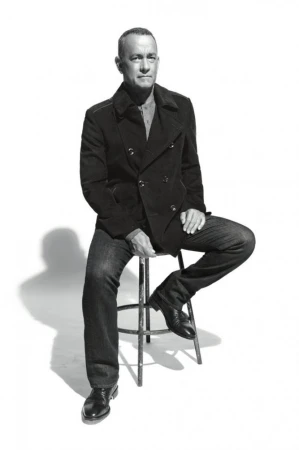 Black and white portrait of adult male in peacoat and jeans, sitting on metal stool in photo studio, looking directly at camera.