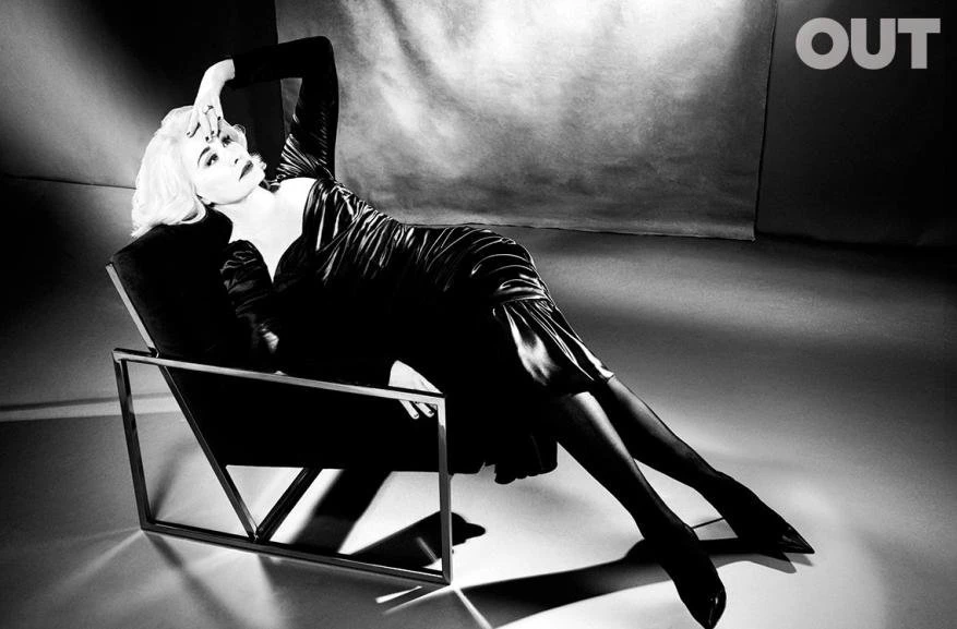 Monochrome portrait of a reclining woman with platinum hair wearing a black dress and stockings against a neutral background.
