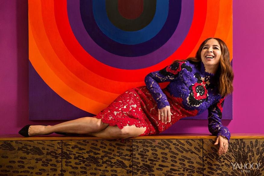 Adult woman in a purple and red lace dress lying on a wooden cabinet with a rainbow circle painting on the wall behind her.