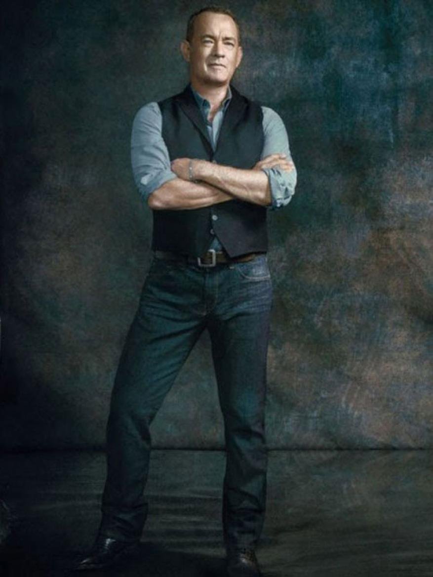Full-length portrait of adult man with crossed arms, wearing dark vest, blue jeans, and black shoes against dark textured backdrop.