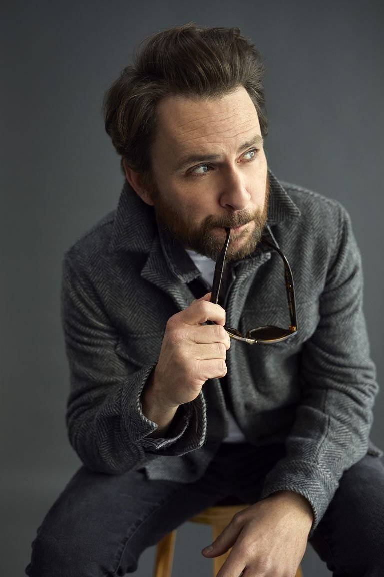 Portrait of a man with a beard and dark hair holding glasses, wearing a herringbone jacket, sitting on a stool in a studio.