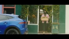 Adult female exits a storefront carrying a basket, parked blue Jaguar E-Pace nearby, set in a daytime outdoor environment.