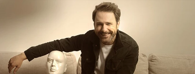 Smiling man wearing a dark jacket over a white t-shirt resting his arm on a white mannequin head next to a sofa
