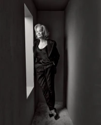 Monochrome portrait of an elderly woman in a dark blazer and trousers posing inside a narrow architectural corridor.