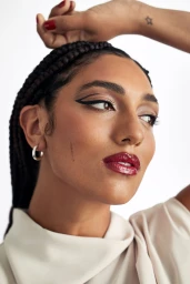 Close-up portrait of a woman with braided dark hair, graphic eyeliner, glossy burgundy lips, and a small silver hoop earring.