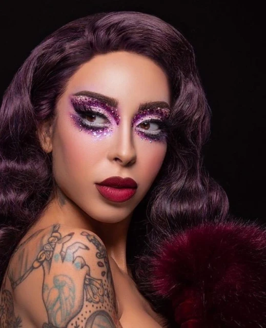 Close-up portrait of a woman with purple hair, glitter eyeshadow, and tattoos on her arm posing against a black background.