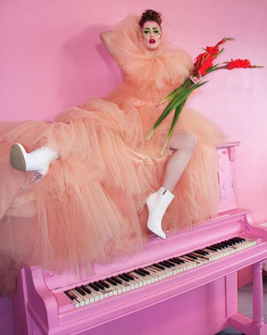 Woman in a peach tulle dress poses with red flowers on a pink piano against a pink backdrop in a fashion photo shoot.