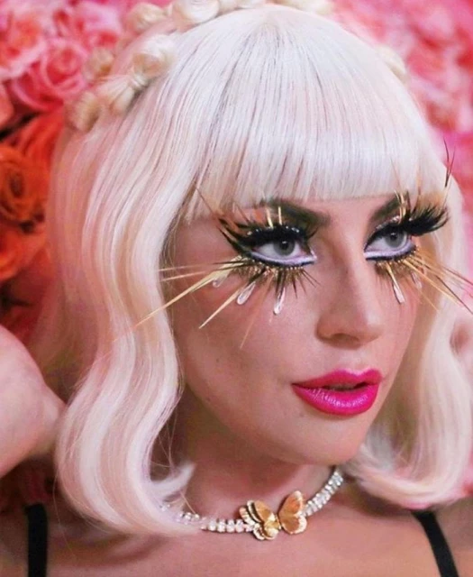 Close-up of a woman with blonde hair, vibrant pink lipstick, extravagant eyelashes, and a butterfly necklace against a floral background.