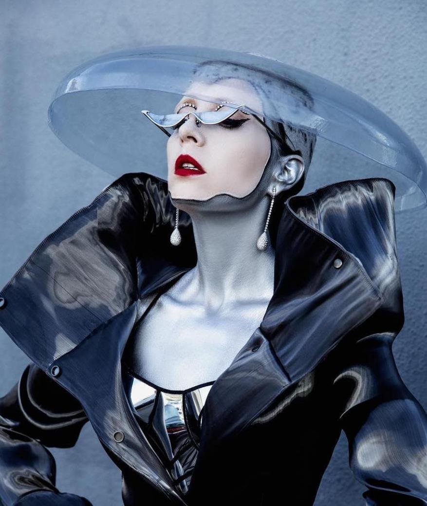 Model showcases a futuristic fashion ensemble with clear hat, dramatic makeup, and a shiny black coat against a neutral background.