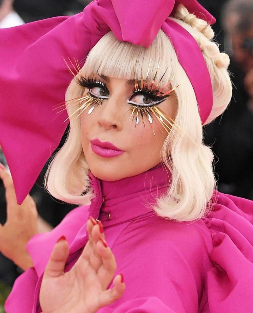 Close-up portrait of a woman with blond hair, pink lipstick, and dramatic gold and black eye makeup wearing a pink bow headband.