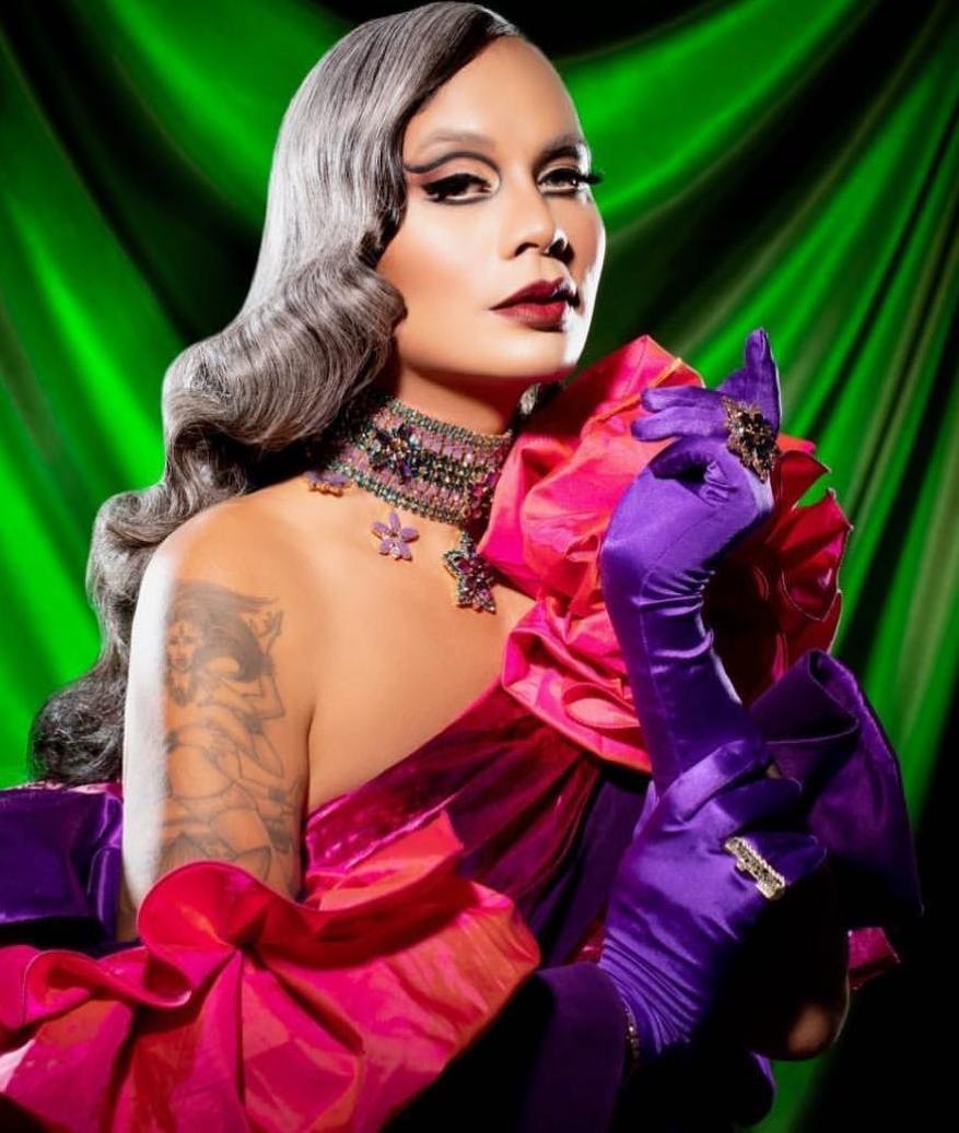 Portrait of a person with grey curly hair wearing heavy makeup and an elaborate pink and purple gown with matching gloves in a studio.