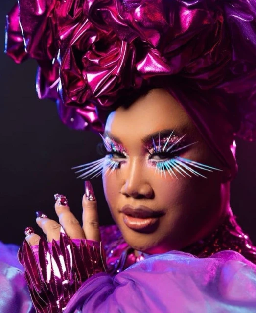 Close-up of a person in elaborate purple headwear and matching makeup, featuring exaggerated eyelashes and manicured nails against a dark background.