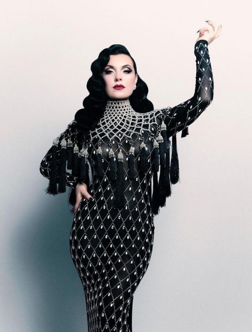 Elegant adult female wearing a floor-length formal gown with black beaded details, dark lipstick, and vintage-style hair, posing in a studio.