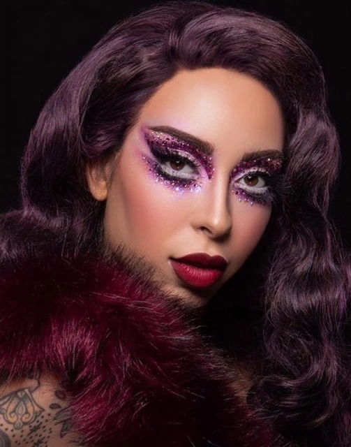 A woman with dramatic purple glitter makeup poses in front of a black background with a purple fur stole draped around her shoulders.