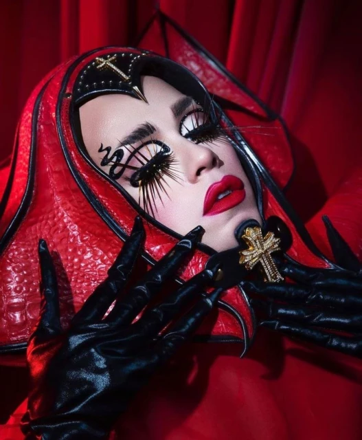 Close-up of a woman with dramatic makeup, long lashes, and a decorative red and black headdress, wearing black gloves and red lipstick.