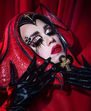 Close-up of a woman with dramatic makeup, long lashes, and a decorative red and black headdress, wearing black gloves and red lipstick.