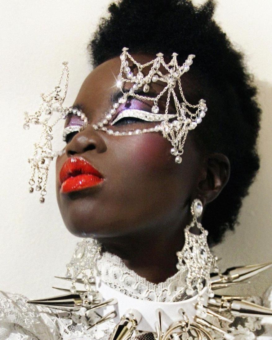 Close-up portrait of a woman with afro, wearing a silver jeweled mask, spike collar necklace, statement earrings, and vibrant red lipstick.