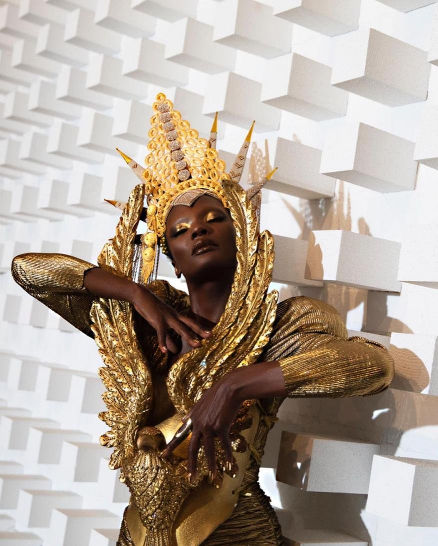 Stunning portrait of a black woman with gold eye makeup, gold headdress and gold feather dress against a textured white background.