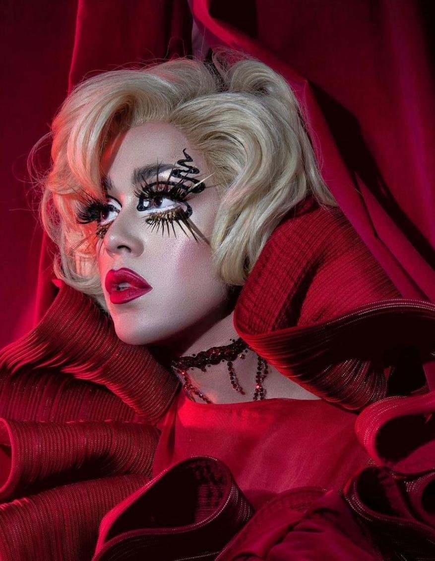 Blonde haired person in elaborate eye makeup, red lipstick, and frilly red dress with a red curtain in the background.