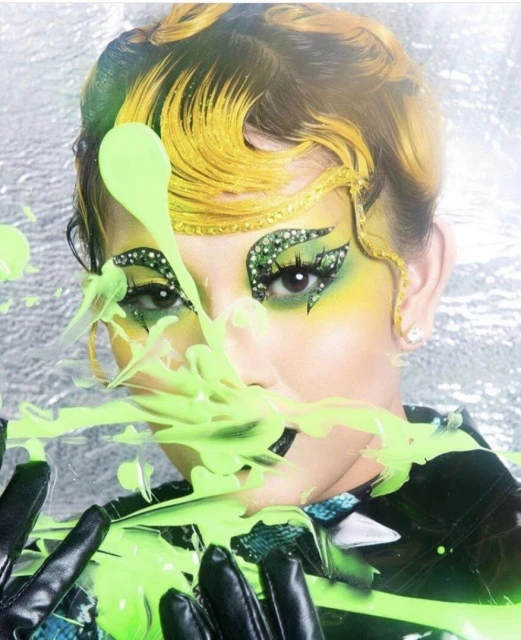 Close-up of a person with dramatic makeup including neon green and rhinestones with some green paint spilling across their face.