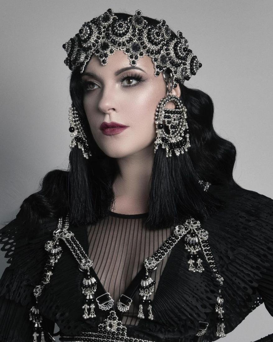 Woman wearing a detailed black beaded headdress and shoulder piece poses in a studio. The headdress has an elaborate beaded design.