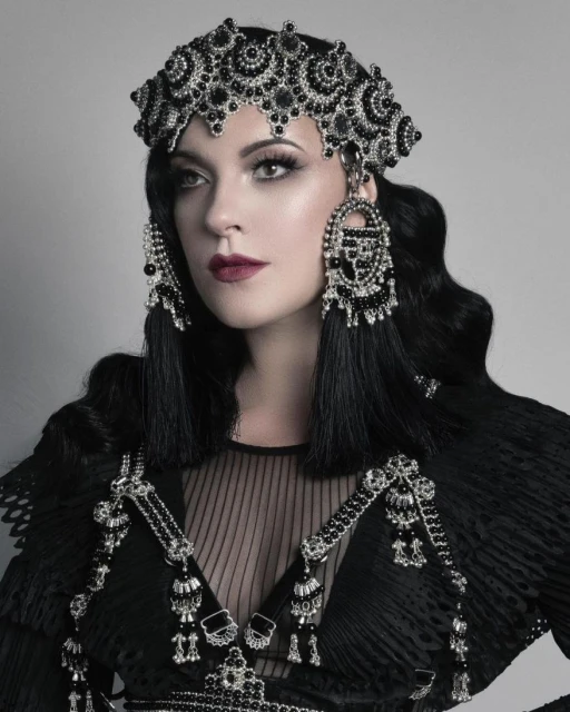 Woman wearing a detailed black beaded headdress and shoulder piece poses in a studio. The headdress has an elaborate beaded design.