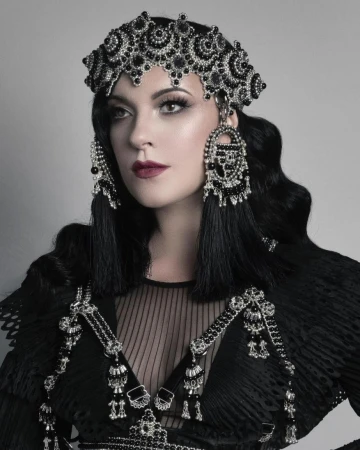 Woman wearing a detailed black beaded headdress and shoulder piece poses in a studio. The headdress has an elaborate beaded design.