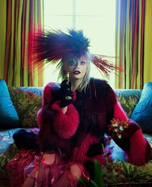 Portrait of a woman wearing a colorful fur coat and feathered hat, peering through a magnifying glass, seated on a floral sofa.