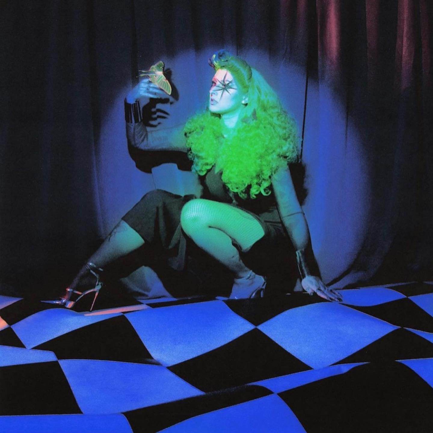 Woman in a dark dress with green hair and a spider face tattoo sits cross-legged on a black-and-blue checkerboard pattern with a moth in hand.