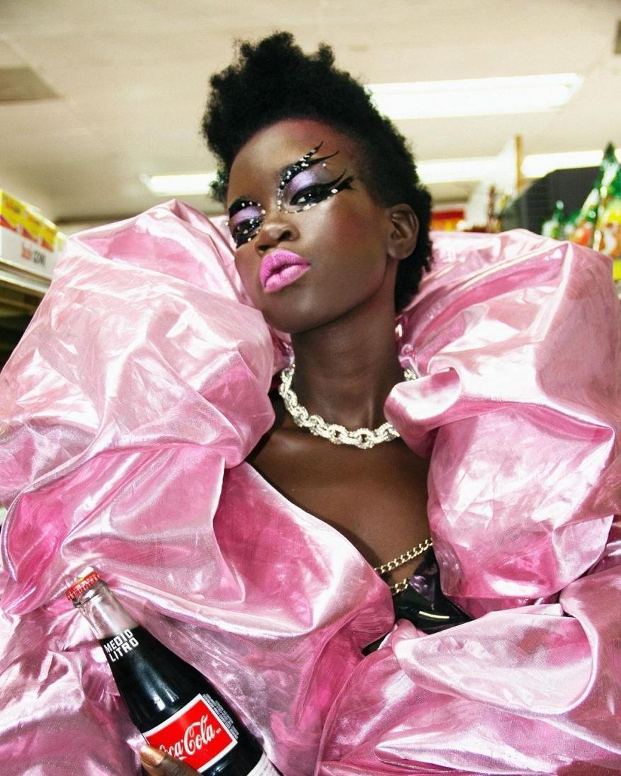Glamorous woman in avant-garde pink ruffled gown with Coca-Cola bottle in a grocery setting, showcasing striking makeup and unique style.