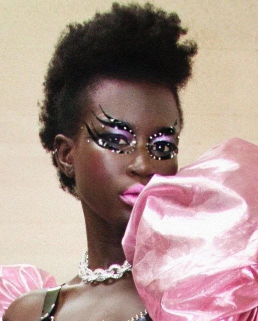 Dark-skinned woman with distinctive eye makeup and a pink, oversized puff sleeve. She wears a silver chain and has dark, curly hair.