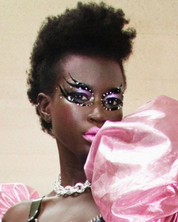 Dark-skinned woman with distinctive eye makeup and a pink, oversized puff sleeve. She wears a silver chain and has dark, curly hair.
