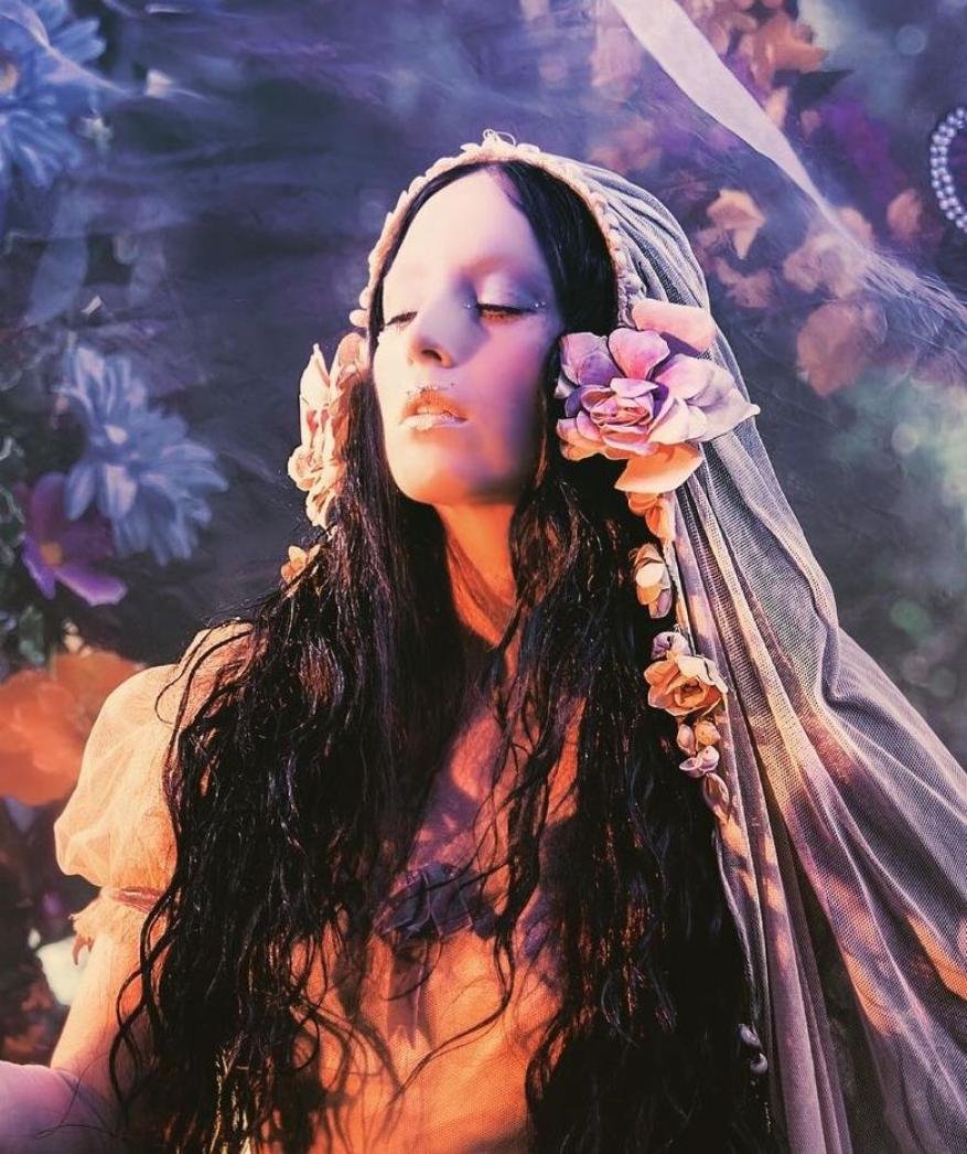 Dreamy portrait of a woman with closed eyes and long, dark wavy hair, adorned with flowers and a sheer head covering, floral background.