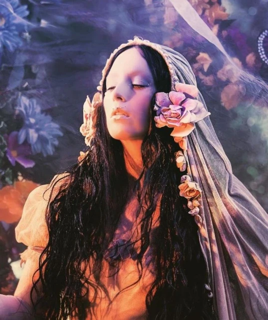 Dreamy portrait of a woman with closed eyes and long, dark wavy hair, adorned with flowers and a sheer head covering, floral background.