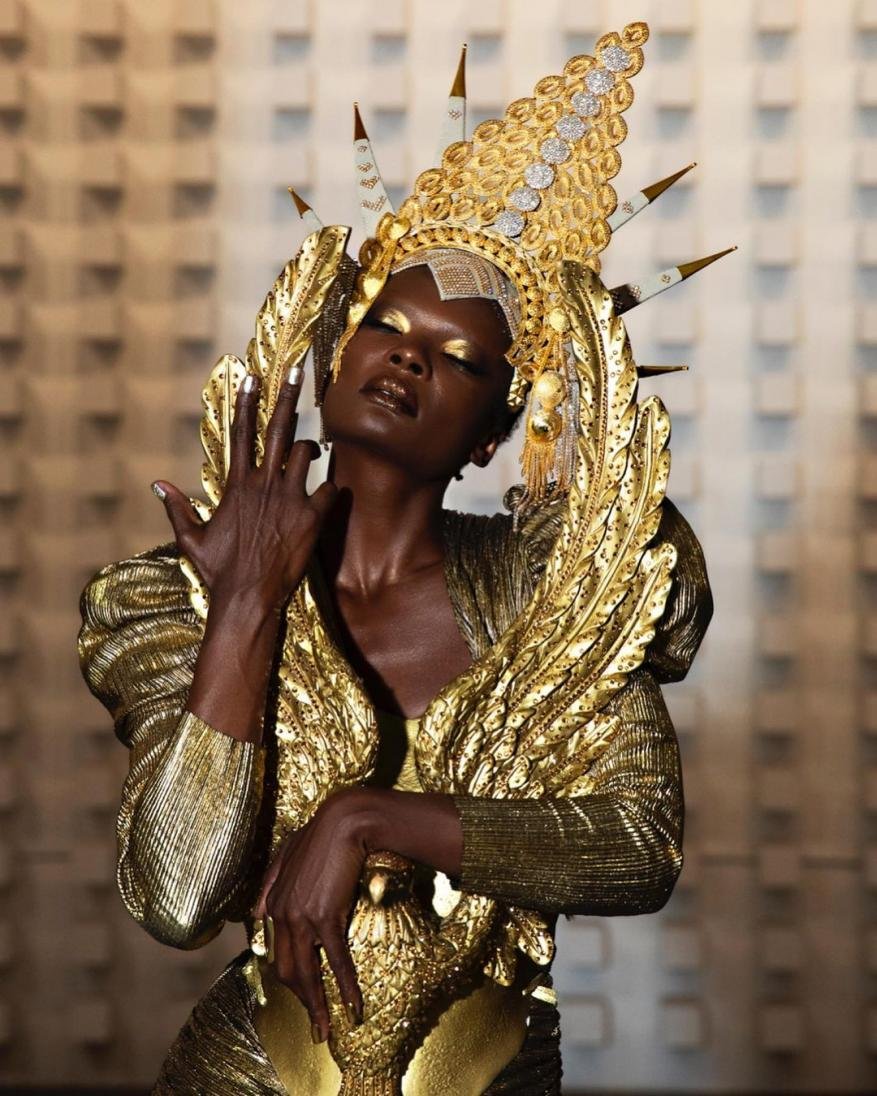 Elegant African woman in elaborate gold costume and headpiece poses against a geometric wall, showcasing beauty and artistic design.