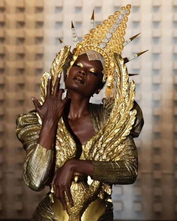 Elegant African woman in elaborate gold costume and headpiece poses against a geometric wall, showcasing beauty and artistic design.