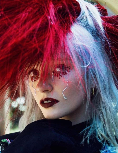 Close-up portrait of a woman with red and white hair, dark lipstick, and face gems, posed in front of a blurred outdoor background.